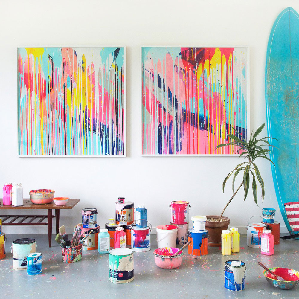 A collection of vibrant, abstract painting prints displayed in an artist's studio setting with paint containers and a surfboard in the foreground.